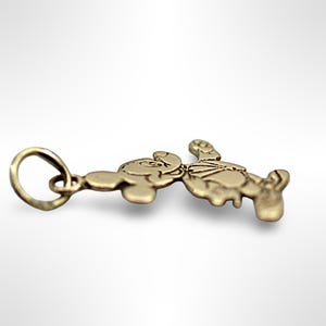 Puede incluir: Un dije de oro con un personaje de dibujos animados, Mickey Mouse, en una pose de pie con los brazos levantados. El dije tiene un bucle para sujetarlo a un collar o pulsera.