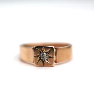 May include: A rose gold ring featuring a square-shaped centerpiece with a starburst engraving. A single, small, clear, round-cut gemstone is set in the center of the starburst design. The band is smooth and solid, maintaining a consistent width.
