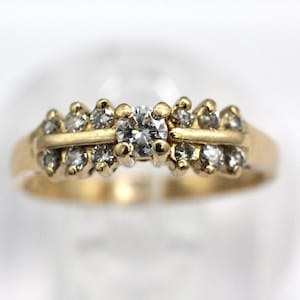 May include: A gold ring with a central diamond and smaller diamonds on either side. The ring has a classic design with a polished finish, showcasing the sparkle of the gemstones. The ring is set on a white background.