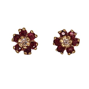 May include: A pair of gold-colored flower stud earrings. Each earring features a central, round, clear stone surrounded by six deep red gemstones. The earrings are set against a white background.