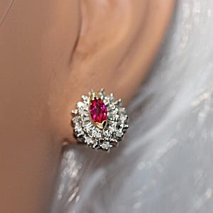 May include: A cluster earring featuring a central marquise-cut red gemstone set in yellow gold, surrounded by a double halo of small round white diamonds set in white metal. The earring has a circular shape and a high-shine metallic finish.