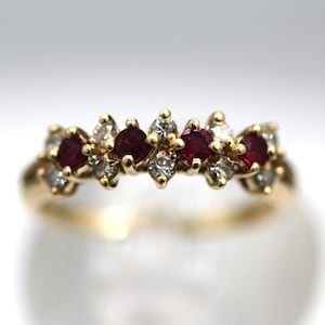 May include: A gold band ring featuring a row of alternating round, deep red gemstones and small, clear gemstones. The ring has a classic design with a polished finish, suitable for everyday wear or special occasions.
