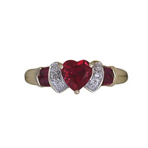 May include: A gold ring featuring a heart-shaped, deep red gemstone as the centerpiece. The ring is flanked by two smaller, rectangular red stones and accented with small, clear gemstones. The band is a polished gold color.