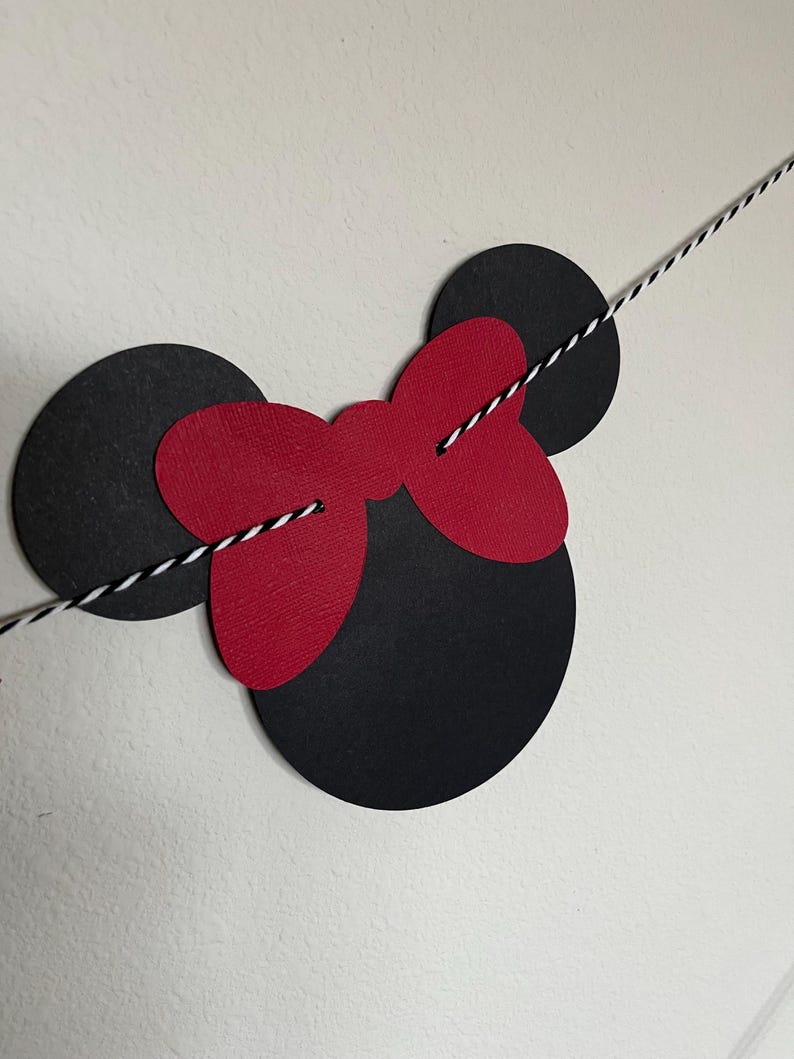 Puede incluir: Recortes de papel negro y rojo de orejas de Mickey Mouse y un lazo, ensartados con un cordel blanco y negro.