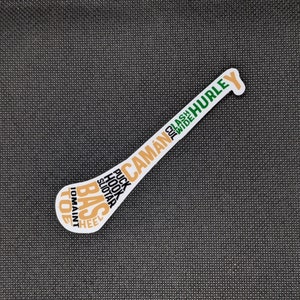 Puede incluir: Una pegatina blanca con forma de palo de hurley, con texto en naranja, verde y blanco. El texto incluye palabras como "CAM&Aacute;N", "HURLEY", "PUCK" y "HOOK". La pegatina est&aacute; sobre una superficie texturizada gris oscuro.