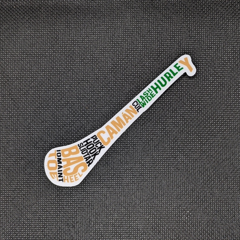 Puede incluir: Una pegatina blanca con forma de palo de hurley, con texto en naranja, verde y blanco. El texto incluye palabras como "CAM&Aacute;N", "HURLEY", "PUCK" y "HOOK". La pegatina est&aacute; sobre una superficie texturizada gris oscuro.