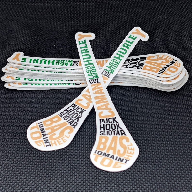 Puede incluir: Una pila de pegatinas blancas con forma de palo de hurley, con texto verde y naranja. El texto incluye "HURLEY", "CAMAN" y "PUCK HOOK SLIOTAR". Las pegatinas est&aacute;n sobre una superficie negra.