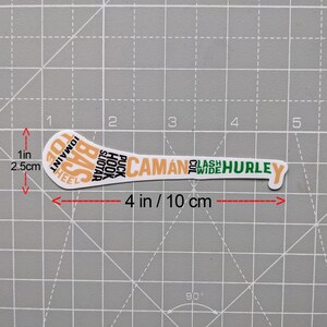 Puede incluir: Una pegatina blanca con forma de palo de hurley, de 10 cm de largo, con texto naranja y verde. El texto incluye "CAM&Aacute;N" y "HURLEY". La pegatina est&aacute; sobre un fondo de cuadr&iacute;cula gris.