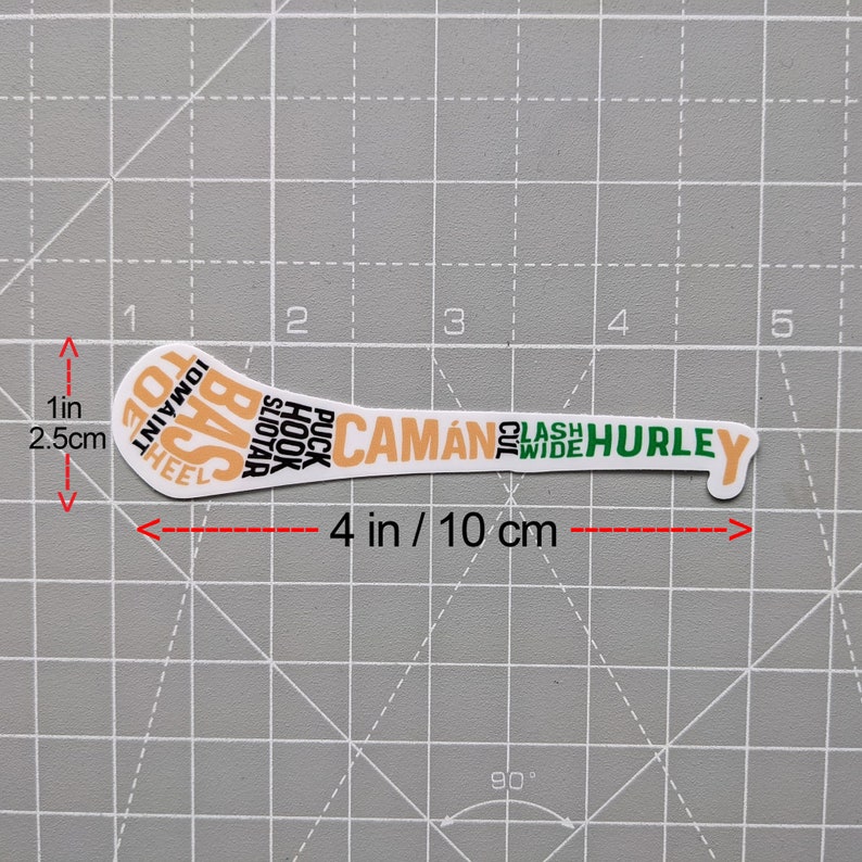 Puede incluir: Una pegatina blanca con forma de palo de hurley, de 10 cm de largo, con texto naranja y verde. El texto incluye "CAM&Aacute;N" y "HURLEY". La pegatina est&aacute; sobre un fondo de cuadr&iacute;cula gris.
