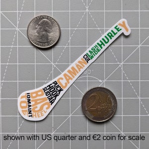 Puede incluir: Una pegatina blanca con forma de palo de hockey, con texto en naranja, verde y negro. El texto incluye "CAM&Aacute;N", "HURLEY", "PUCK", "HOOK" y "SLIOTAR". Un cuarto de d&oacute;lar estadounidense y una moneda de 2 &euro; est&aacute;n al lado.