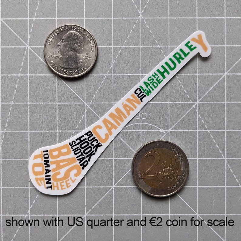 Puede incluir: Una pegatina blanca con forma de palo de hockey, con texto en naranja, verde y negro. El texto incluye "CAM&Aacute;N", "HURLEY", "PUCK", "HOOK" y "SLIOTAR". Un cuarto de d&oacute;lar estadounidense y una moneda de 2 &euro; est&aacute;n al lado.