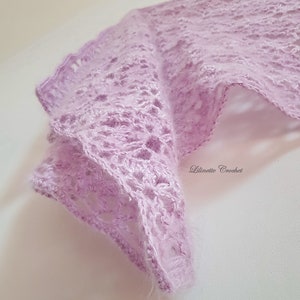 Puede incluir: Un chal delicado de color lila claro tejido a crochet con un patrón de encaje. El chal está hecho con un hilo suave y peludo y tiene una textura fluida y drapeada.