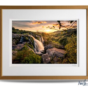 Puede incluir: Una fotograf&iacute;a de una cascada que cae sobre rocas con una puesta de sol en el fondo. La imagen est&aacute; enmarcada con un marco de color dorado y tiene el texto "Llyn y Fan Fach in Golden Light at Sunset" y una firma en la parte inferior.