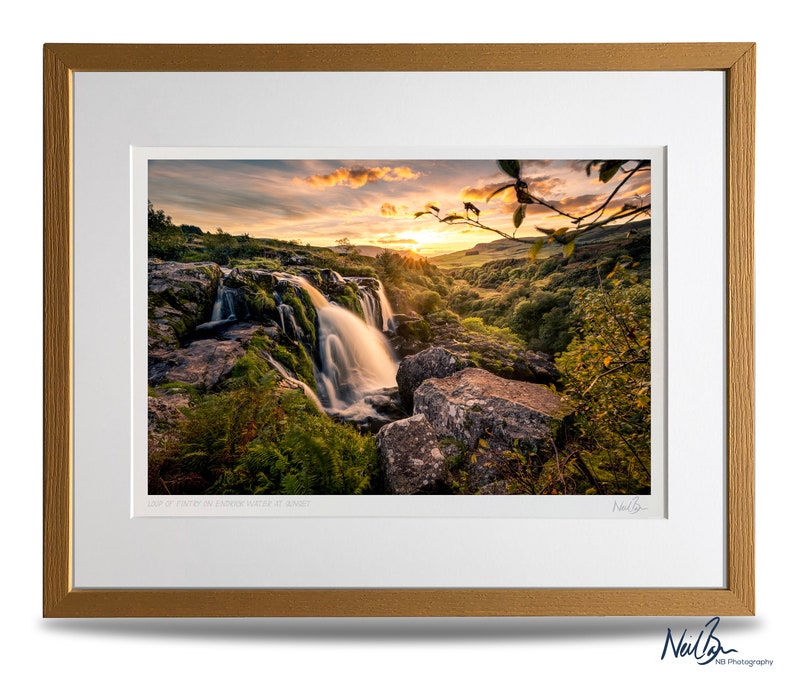 Puede incluir: Una fotograf&iacute;a de una cascada que cae sobre rocas con una puesta de sol en el fondo. La imagen est&aacute; enmarcada con un marco de color dorado y tiene el texto "Llyn y Fan Fach in Golden Light at Sunset" y una firma en la parte inferior.
