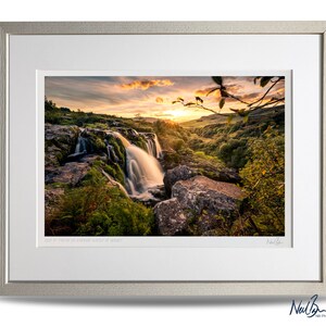 Puede incluir: Una fotograf&iacute;a enmarcada de una cascada que cae sobre rocas cubiertas de musgo, con un cielo de puesta de sol vibrante en el fondo. La imagen se titula "Sun at Forty Foot in Cwmduad Water at Sunset".