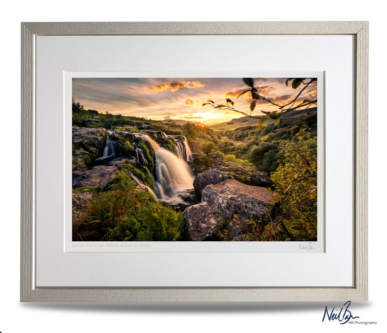 Puede incluir: Una fotograf&iacute;a enmarcada de una cascada que cae sobre rocas cubiertas de musgo, con un cielo de puesta de sol vibrante en el fondo. La imagen se titula "Sun at Forty Foot in Cwmduad Water at Sunset".