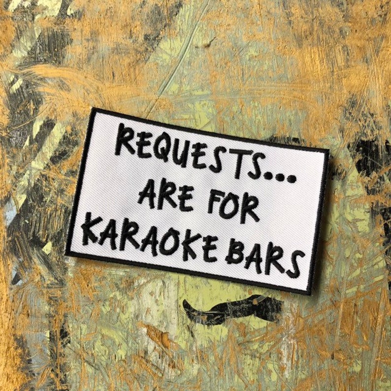 K&ouml;nnte beinhalten: Wei&szlig;er Stoff-Patch mit schwarzem gesticktem Text, der "Requests... Are for Karaoke Bars" lautet.