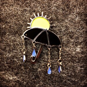 May include: A black umbrella with a yellow sun on top and gold chains with blue raindrops hanging from the bottom.
