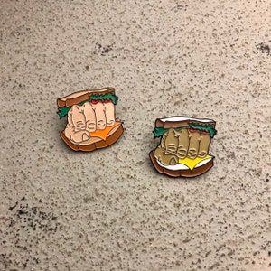 May include: Two enamel pins depicting a hand clenching a sandwich. The sandwich on the left has a brown hand and the sandwich on the right has a tan hand.
