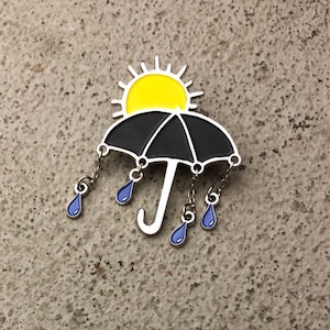 May include: A black umbrella with a yellow sun on top and blue raindrops hanging from the bottom. The umbrella is a metal pin.