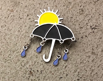 May include: A black umbrella with a yellow sun on top and blue raindrops hanging from the bottom. The umbrella is a metal pin.