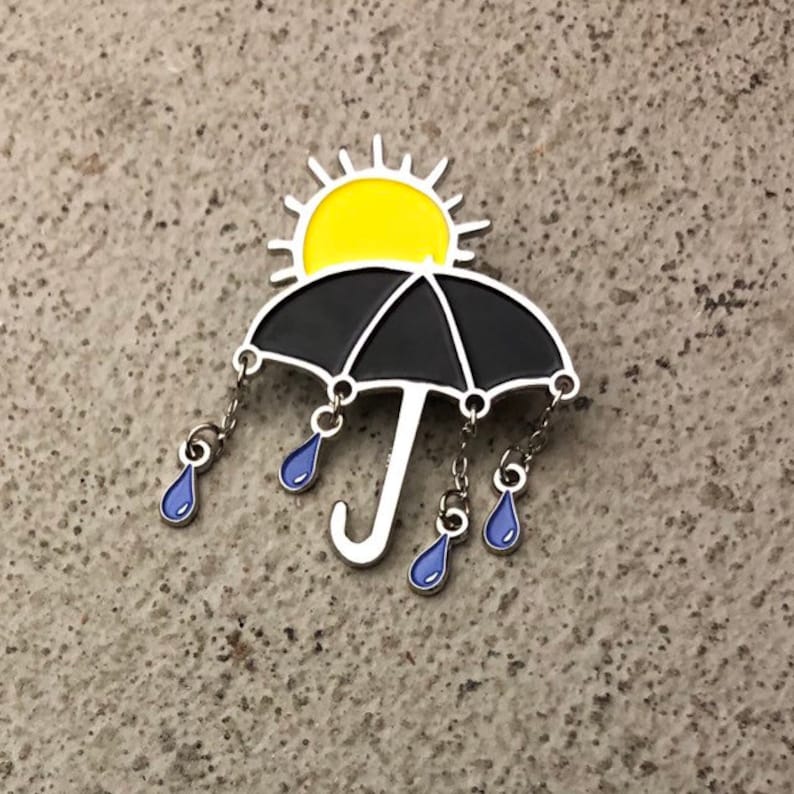 May include: A black umbrella with a yellow sun on top and blue raindrops hanging from the bottom. The umbrella is a metal pin.