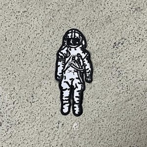 Op de afbeelding: Zwart-witte geborduurde patch van een astronaut in een ruimtepak. De astronaut staat met zijn armen langs zijn zijde.
