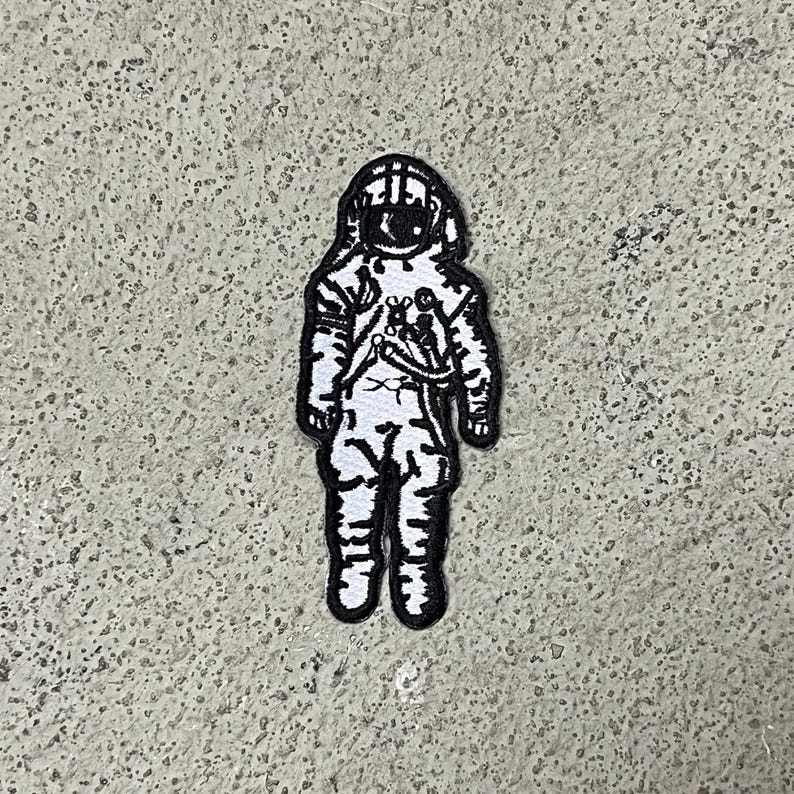 Op de afbeelding: Zwart-witte geborduurde patch van een astronaut in een ruimtepak. De astronaut staat met zijn armen langs zijn zijde.