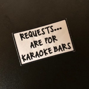 K&ouml;nnte beinhalten: Wei&szlig;er rechteckiger Patch mit schwarzem gesticktem Text, der "REQUESTS... ARE FOR KARAOKE BARS" lautet.