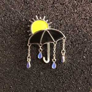 May include: A black umbrella with a yellow sun on top and blue raindrops hanging from chains. The umbrella is a metal pin with enamel details.