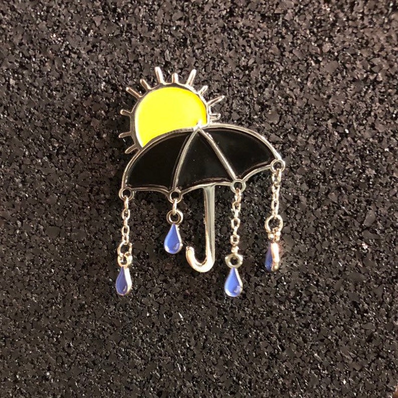 May include: A black umbrella with a yellow sun on top and blue raindrops hanging from chains. The umbrella is a metal pin with enamel details.