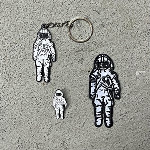 Op de afbeelding: Drie astronaut-patches, waarvan er &eacute;&eacute;n een sleutelhanger heeft. De patches zijn zwart-wit met een eenvoudig ontwerp. De astronaut draagt een helm en een ruimtepak.
