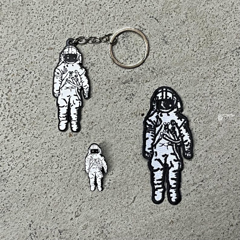 Op de afbeelding: Drie astronaut-patches, waarvan er &eacute;&eacute;n een sleutelhanger heeft. De patches zijn zwart-wit met een eenvoudig ontwerp. De astronaut draagt een helm en een ruimtepak.