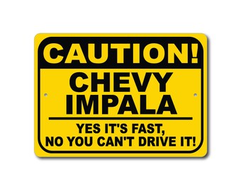 May include: A yellow rectangular sign with black text that reads "CAUTION! CHEVY IMPALA YES IT'S FAST, NO YOU CAN'T DRIVE IT!" The sign has rounded corners and two small holes for mounting.