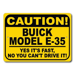 May include: A yellow rectangular sign with black text that reads "CAUTION! BUICK MODEL E-35 YES IT'S FAST, NO YOU CAN'T DRIVE IT!". The sign has rounded corners and two small holes for mounting.