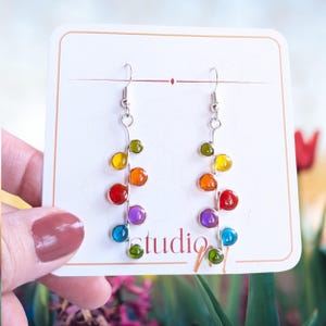 May include: Dangle earrings featuring a silver-toned wire stem adorned with small, round, multi-colored glass beads in shades of green, yellow, orange, red, purple, and blue. The earrings are displayed on a white card with the text studio.