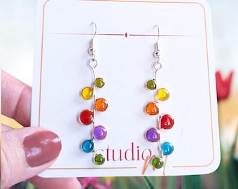 May include: Dangle earrings featuring a silver-toned wire stem adorned with small, round, multi-coloured glass beads in shades of green, yellow, orange, red, purple, and blue. The earrings are displayed on a white card with the text studio.