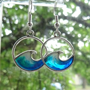 May include: A pair of silver-toned dangle earrings featuring a circular frame enclosing a stylized ocean wave design. The wave is filled with a translucent, swirling gradient of deep blue, turquoise, and white resin, suspended from simple silver hooks.