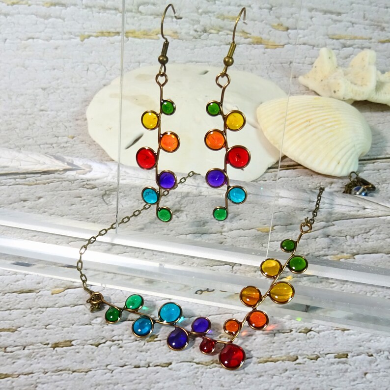 May include: A rainbow colored glass bead necklace and matching earrings. The necklace and earrings are made of small, round glass beads that are strung on a wire. The beads are arranged in a pattern that resembles a vine.