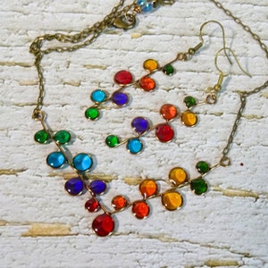 May include: A rainbow colored glass bead necklace and matching earrings. The necklace and earrings are made of small, round glass beads that are strung on a wire. The beads are arranged in a pattern that resembles a branch of leaves. The necklace is attached to a brass chain.