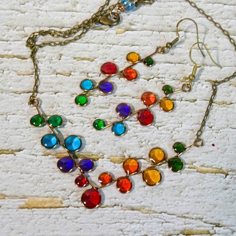 May include: A rainbow colored glass bead necklace and matching earrings. The necklace and earrings are made of small, round glass beads that are strung on a wire. The beads are arranged in a pattern that resembles a branch of leaves. The necklace is attached to a brass chain.