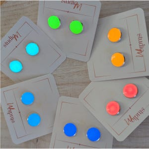 May include: A collection of colorful stud earrings in neon green, orange, blue, and pink. Each pair is displayed on a white card with the text "studioM" in a handwritten font. The earrings have a metallic silver border.