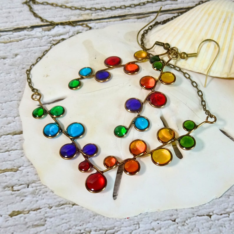 May include: A rainbow colored glass bead necklace and earrings set. The necklace and earrings are made of small, round glass beads that are strung on a wire. The beads are arranged in a rainbow pattern, with each bead a different color. The necklace is long and has a simple clasp. The earrings are small and dangle from small hoops.