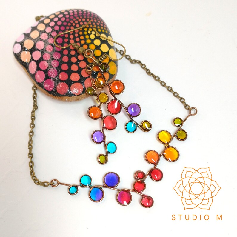May include: A rainbow colored glass bead necklace and earrings with a delicate wire design. The necklace and earrings are made with small, round glass beads in a variety of colors, including red, orange, yellow, green, blue, and purple. The beads are strung on a thin wire that is twisted into a delicate vine-like design. The necklace is long and has a clasp closure. The earrings are dangle earrings and have a small hook closure.