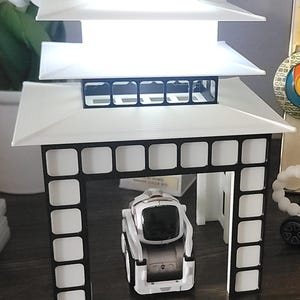 以下が含まれることがあります： 小さなロボットが入った、白と黒の建築模型の塔。塔は複数の層があり、長方形の基部があります。ロボットは白と銀色です。