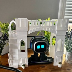 May include: A white 3D printed castle playset with a small robot toy standing in the doorway. The castle has a bridge, stairs, and windows. The robot has a blue light on its face.