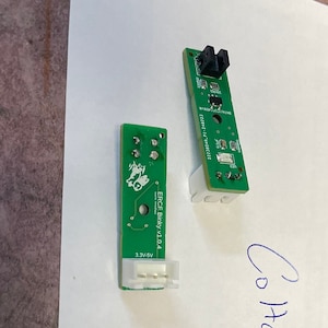 May include: Two green circuit boards with black components. The larger board has the text "EBC1 Display V1.04" and "3.3V only". The smaller board has a black sensor on the top. The text "Co-Hen V2.229 V2.6.1991" is written on the white paper below the boards.