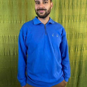 Op de afbeelding: Een felblauw sweatshirt met lange mouwen en een kraag met een kwart rits. Het sweatshirt heeft een klein, wit geborduurd ontwerp op de borst. De persoon op de foto draagt een bruine broek. De achtergrond is een gestructureerde, olijfgroene stof.