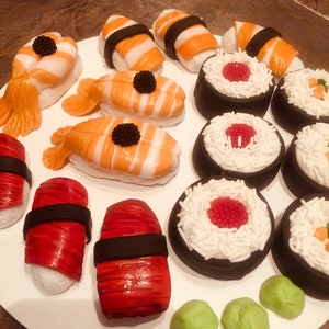 以下が含まれることがあります： 白いプレートに、寿司をテーマにしたケーキポップが盛り付けられています。ケーキポップは、白、オレンジ、黒、赤のフォンダンで飾られています。緑のワサビのケーキポップもあります。