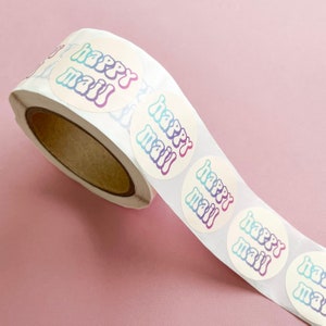 May include: A roll of white round stickers with the text "happy mail" in a rainbow gradient. The stickers are on a pink background.